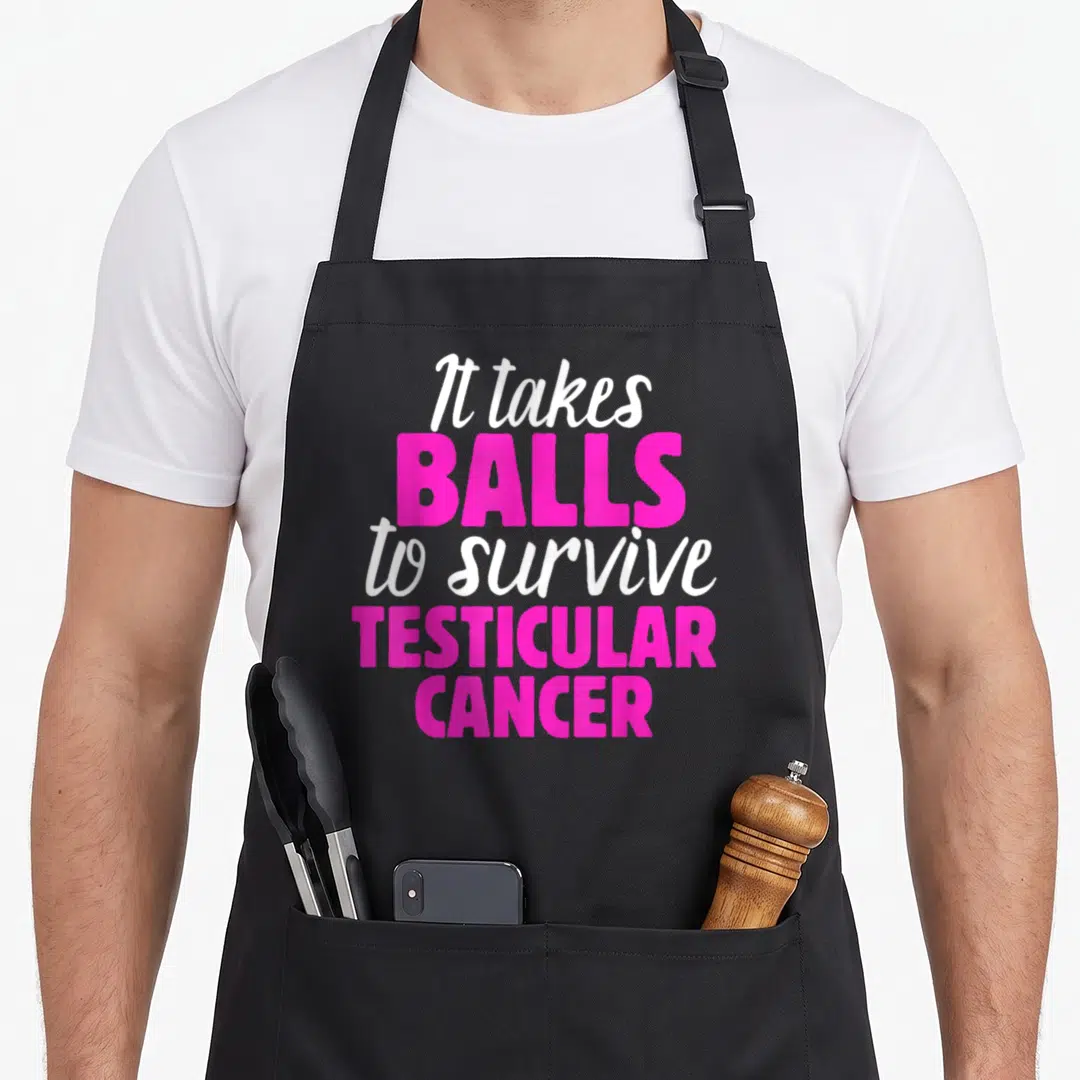 Testicular funny warrior apron - It takes balls to survive testicular cancer text in white and pink stacked typography — shown on a black cotton canvas apron against a white background