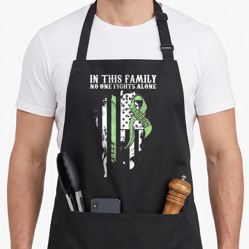 Celiac ribbon apron - Distressed flag with green awareness ribbon containing warrior text and family slogan — shown on a black cotton canvas apron against a white background