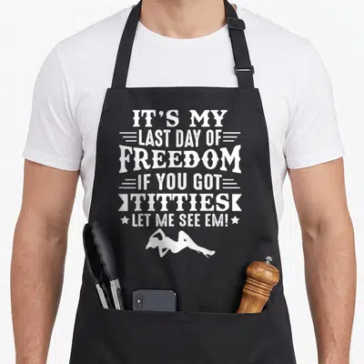 Stag gag aprons - White stacked text IT'S MY LAST DAY OF FREEDOM with a reclining woman silhouette below — shown on a black cotton canvas apron against a white background