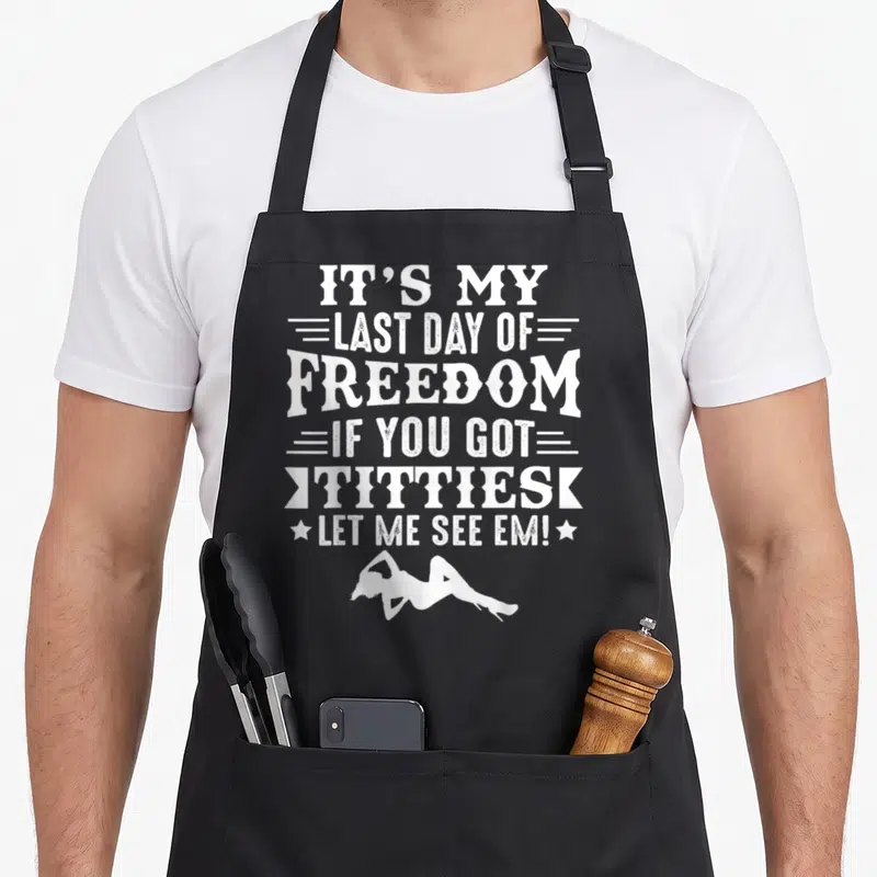 Stag gag aprons - White stacked text IT'S MY LAST DAY OF FREEDOM with a reclining woman silhouette below — shown on a black cotton canvas apron against a white background