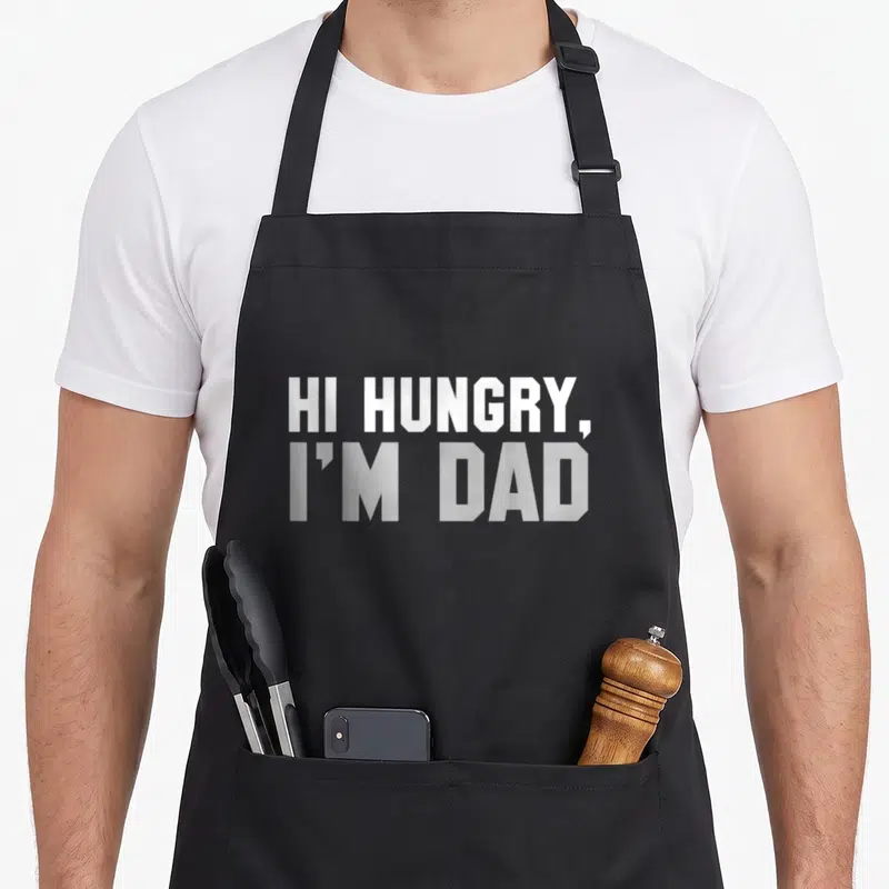 Hungry joke apron - White stacked text reading HI HUNGRY I'M DAD on grey background — shown on a black cotton canvas apron against a white background