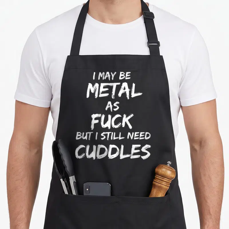 Metalhead heavy metal apron - White brushstroke text reading I may be metal as fuck but I still need cuddles — shown on a black cotton canvas apron against a white background