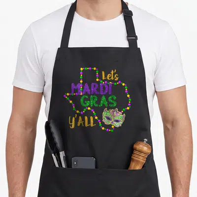 Galveston mardi gras aprons - Let's MARDI GRAS y'all text with masquerade mask and purple green gold bead strands — shown on a black cotton canvas apron against a white background
