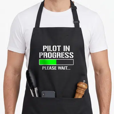 Pilot student aprons - White stencil text reading PILOT IN PROGRESS above a half-filled green progress bar on a black — shown on a black cotton canvas apron against a white background