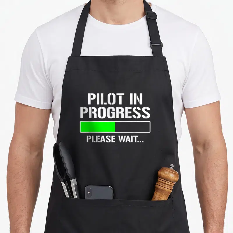 Pilot student aprons - White stencil text reading PILOT IN PROGRESS above a half-filled green progress bar on a black — shown on a black cotton canvas apron against a white background