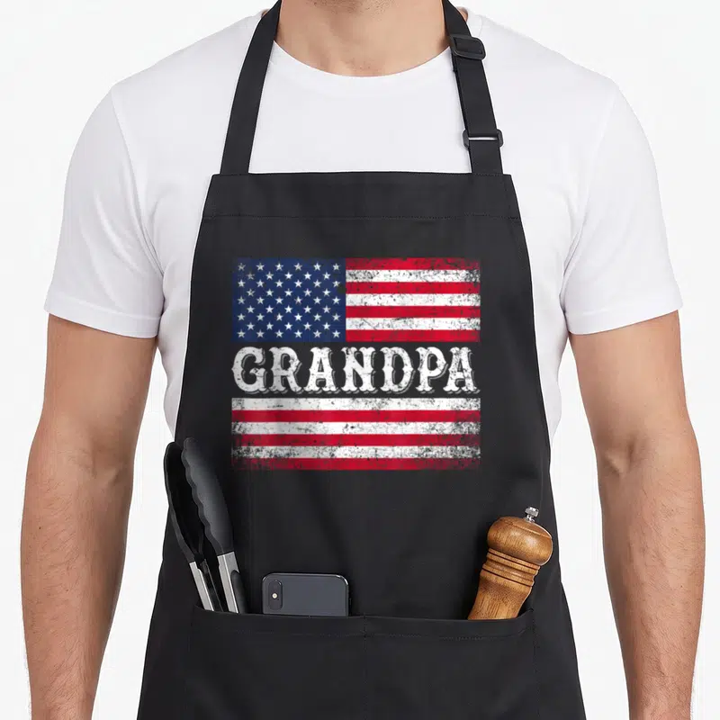 Grandpa flag 4th of july apron - Distressed USA flag with GRANDPA text splitting red and white stripes — shown on a black cotton canvas apron against a white background