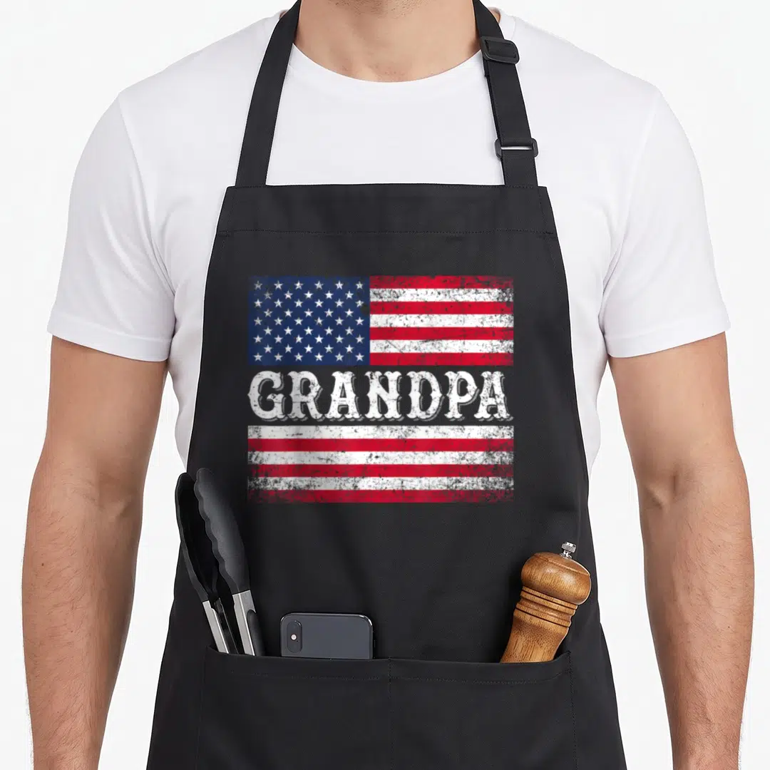 Grandpa flag 4th of july apron - Distressed USA flag with GRANDPA text splitting red and white stripes — shown on a black cotton canvas apron against a white background