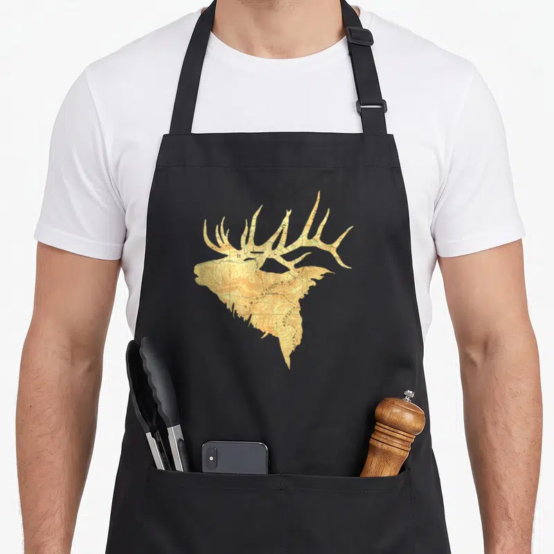 Topographical antler oak apron - Elk head silhouette filled with tan and brown topographic map contour lines — shown on a black cotton canvas apron against a white background