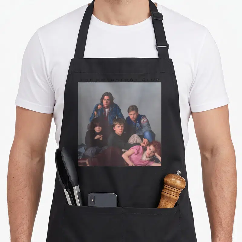 Breakfast century club apron - THE BREAKFAST CLUB text above photo of five teenagers in blue pink green and black — shown on a black cotton canvas apron against a white background
