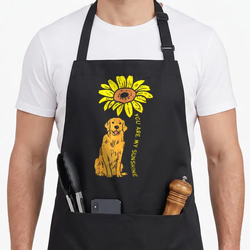 Sunshine golden retriever apron - Cartoon golden retriever sits under yellow sunflower with curved YOU ARE MY SUNSHINE — shown on a black cotton canvas apron against a white background