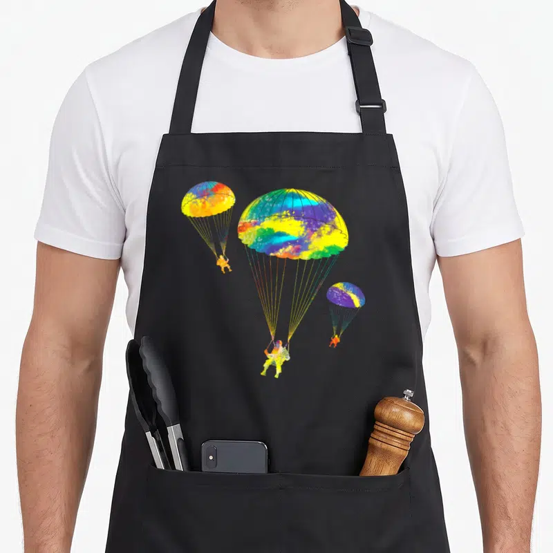 Parachute colorful aprons - Three parachutists with open chutes in watercolor blue yellow red green purple — shown on a black cotton canvas apron against a white background