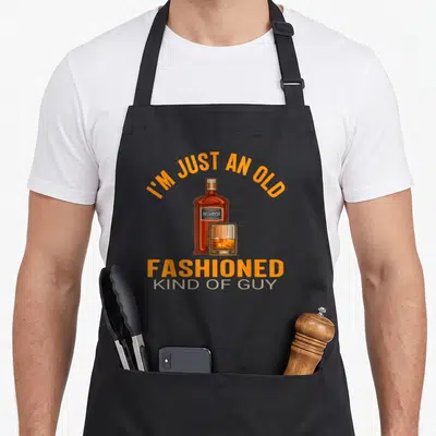 Brandy bourbon old fashioned aprons - Cartoon bourbon bottle and glass with orange arched text on grey background — shown on a black cotton canvas apron against a white background