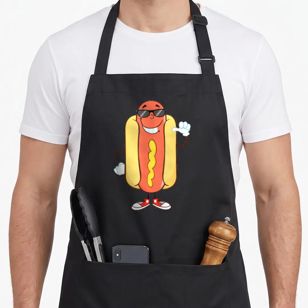 Dancing food aprons - Cartoon hot dog wearing sunglasses and sneakers with yellow mustard line — shown on a black cotton canvas apron against a white background