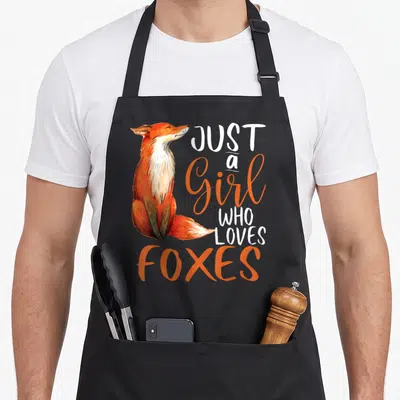 Fox print apron for her - Watercolor red fox sitting next to stacked orange and white text reading JUST a Girl WHO — shown on a black cotton canvas apron against a white background