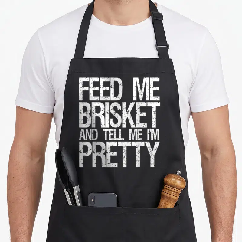 Give texas apron - White stacked text reading FEED ME BRISKET AND TELL ME I'M PRETTY — shown on a black cotton canvas apron against a white background