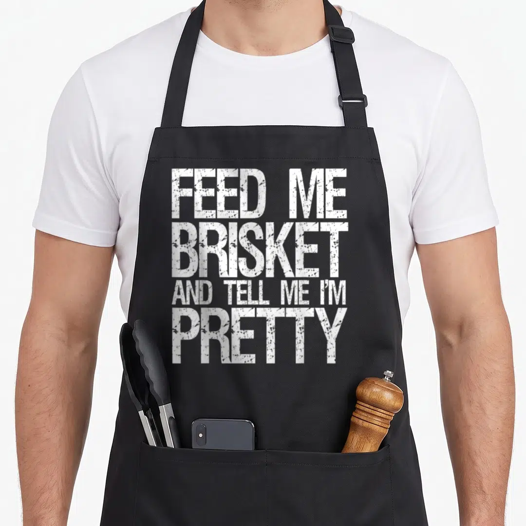 Give texas apron - White stacked text reading FEED ME BRISKET AND TELL ME I'M PRETTY — shown on a black cotton canvas apron against a white background