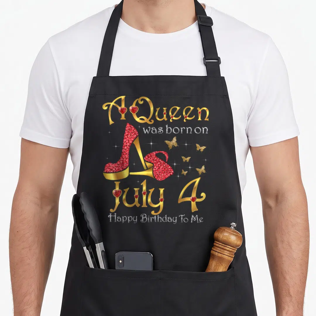 July baking queen apron - A Queen born July 4 text with red high heels, gold butterflies and hearts — shown on a black cotton canvas apron against a white background