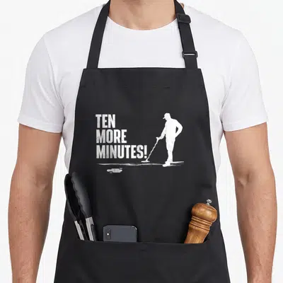 Minute metal detecting apron - Distressed white TEN MORE MINUTES text beside a mopping man silhouette and puddle — shown on a black cotton canvas apron against a white background
