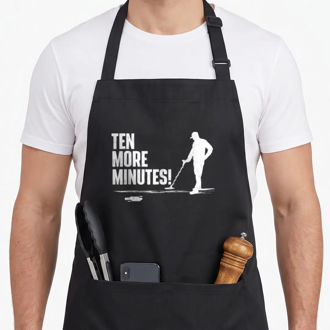 Minute metal detecting apron - Distressed white TEN MORE MINUTES text beside a mopping man silhouette and puddle — shown on a black cotton canvas apron against a white background