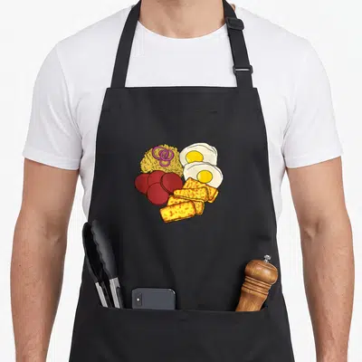Dominican food aprons - Cartoon mangú mound with red salami, fried eggs, and yellow cheese blocks — shown on a black cotton canvas apron against a white background