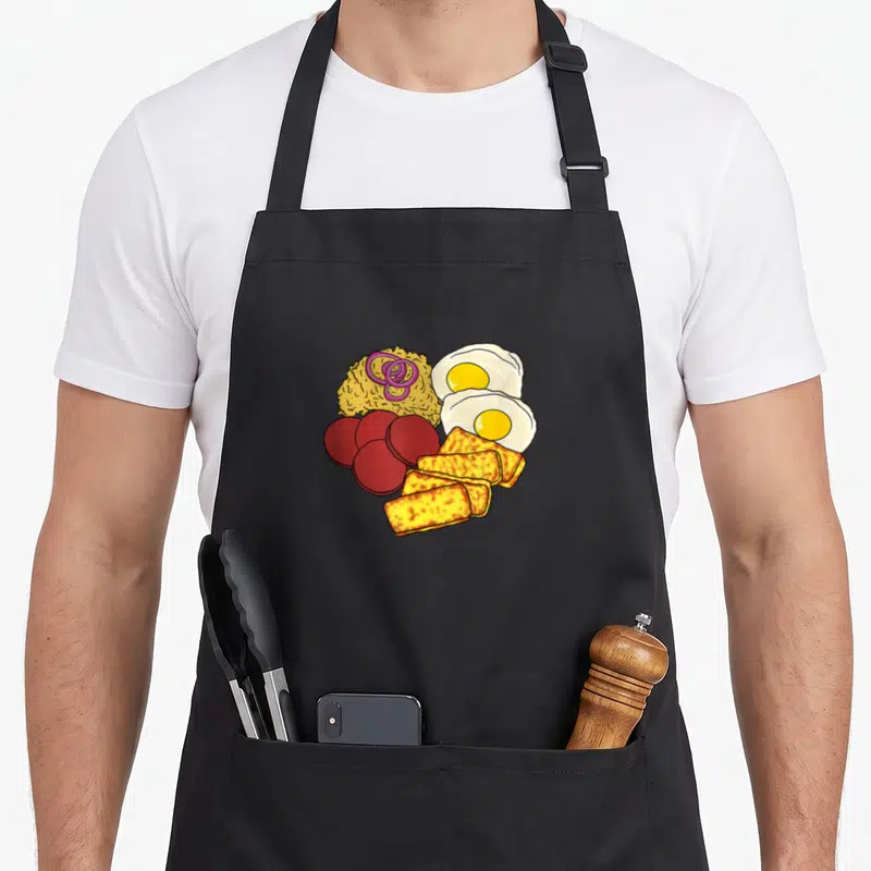 Dominican food aprons - Cartoon mangú mound with red salami, fried eggs, and yellow cheese blocks — shown on a black cotton canvas apron against a white background