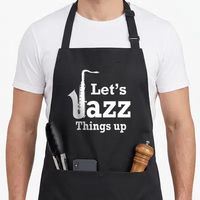 Funny jazz apron - White saxophone graphic overlapping the letter J in stacked Let's Jazz Things up text — shown on a black cotton canvas apron against a white background