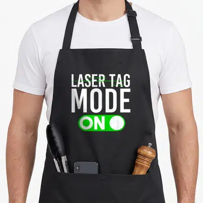 Gun birthday apron - White LASER TAG MODE ON text with green toggle switch graphic on black background — shown on a black cotton canvas apron against a white background