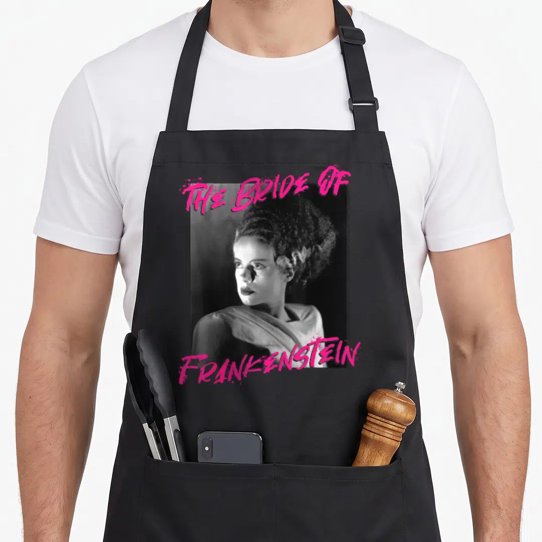 Universal paint splatter apron - Grayscale photo of The Bride of Frankenstein with pink stylized text above and below — shown on a black cotton canvas apron against a white background