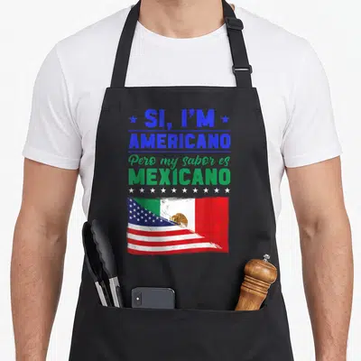 Americano mexican apron - Stacked blue and green text above white stars and a blended USA Mexico flag on black fabric — shown on a black cotton canvas apron against a white background