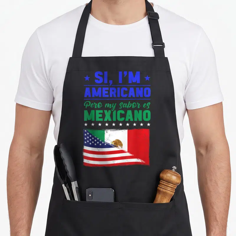 Americano mexican apron - Stacked blue and green text above white stars and a blended USA Mexico flag on black fabric — shown on a black cotton canvas apron against a white background