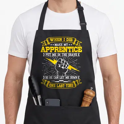 Wiring electrician apron - Retro fist and lightning bolt with ribbon text reading WHEN I DIE MAKE MY APPRENTICE PUT ME — shown on a black cotton canvas apron against a white background