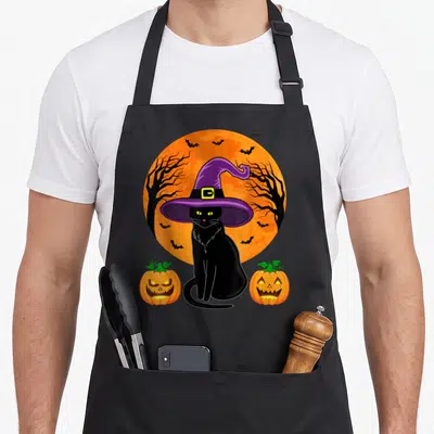 Cat halloween kitchen aprons - Cartoon black cat in purple witch hat between two glowing jack-o'-lanterns against — shown on a black cotton canvas apron against a white background