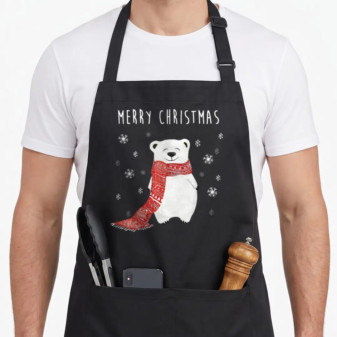 Polar merry christmas apron - Cartoon polar bear in red scarf with white MERRY CHRISTMAS text and snowflakes — shown on a black cotton canvas apron against a white background