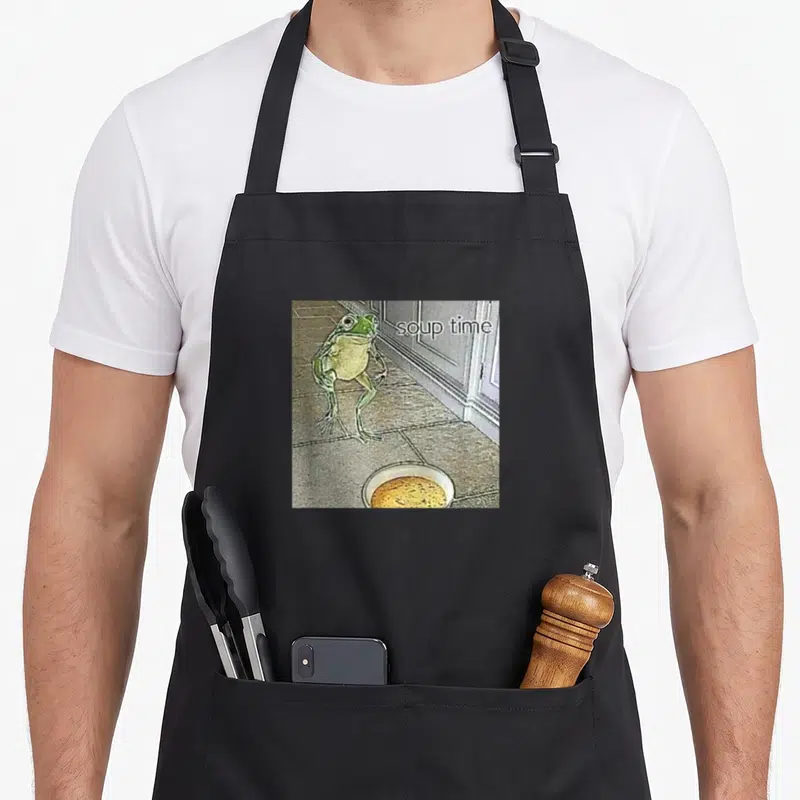 Soup meme apron - Frog standing on tiled floor next to soup bowl with soup time text — shown on a black cotton canvas apron against a white background