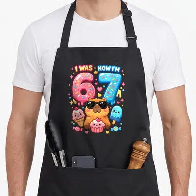Capybara ice cream apron - I WAS NOW I'M text with capybara sunglasses and pink blue numbers — shown on a black cotton canvas apron against a white background