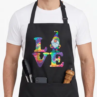 Gnome tie-dye apron - Tie-dye LOVE text with standing hippie gnome replacing the letter O in purple yellow blue red — shown on a black cotton canvas apron against a white background