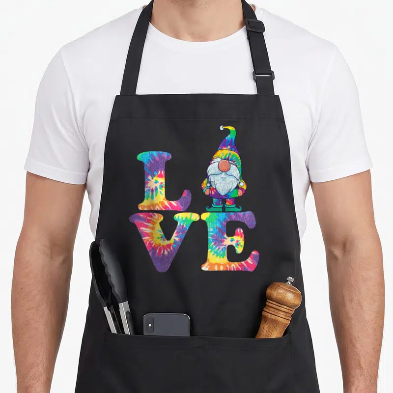 Gnome tie-dye apron - Tie-dye LOVE text with standing hippie gnome replacing the letter O in purple yellow blue red — shown on a black cotton canvas apron against a white background