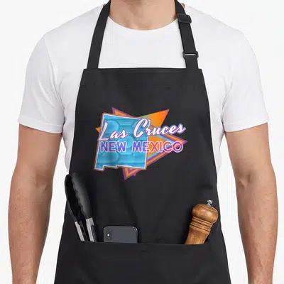Throwback mexico apron - Las Cruces New Mexico text over blue state outline and orange purple triangles — shown on a black cotton canvas apron against a white background
