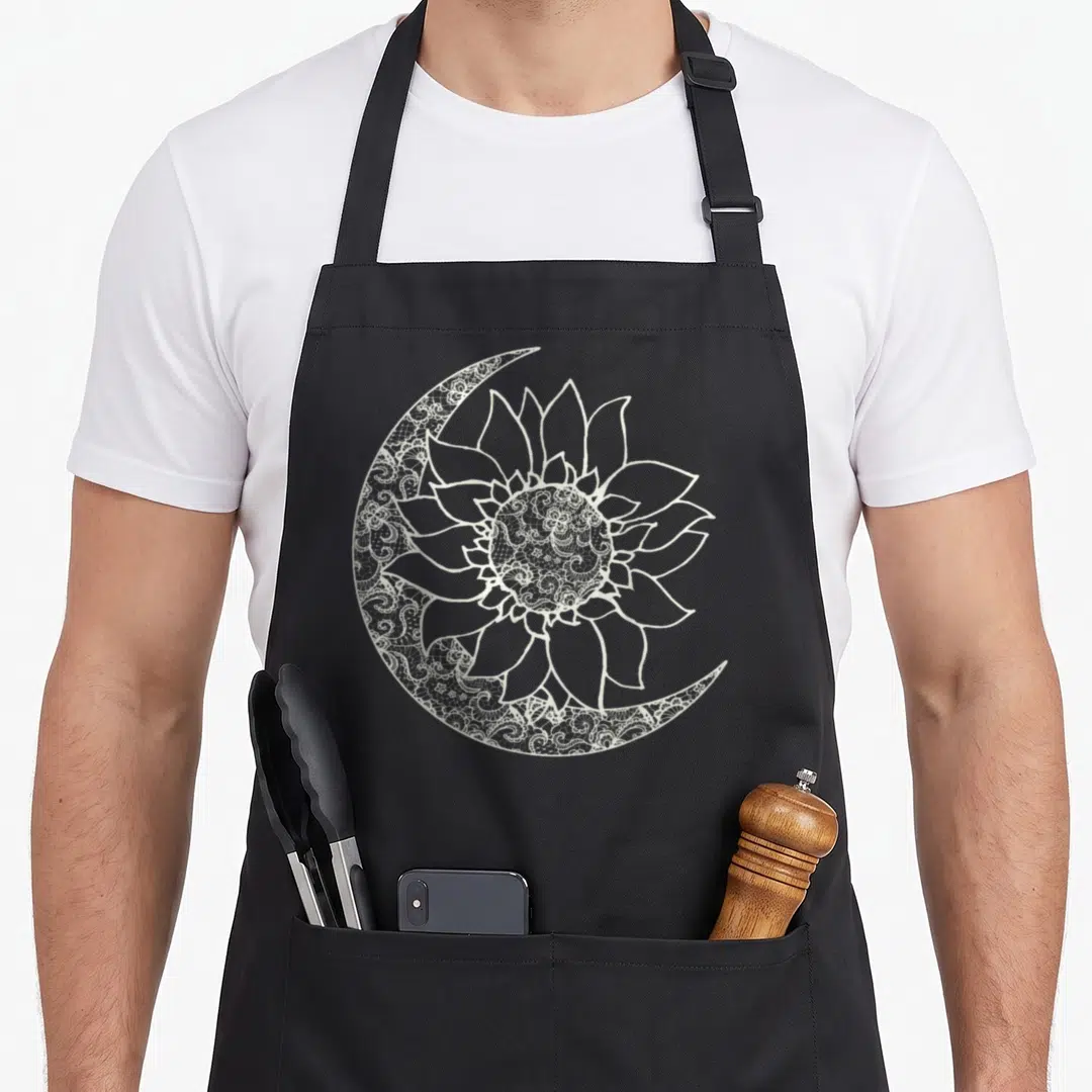 Moon sunflower apron - White line-art sunflower overlapping a lace-filled crescent moon on black fabric — shown on a black cotton canvas apron against a white background
