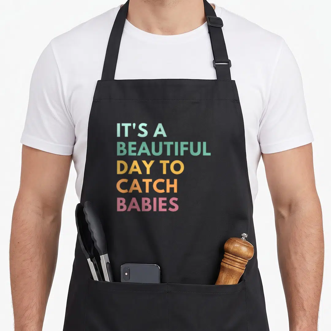 Catch nurse apron - Stacked gradient text IT'S A BEAUTIFUL DAY TO CATCH BABIES in grey green yellow orange pink — shown on a black cotton canvas apron against a white background