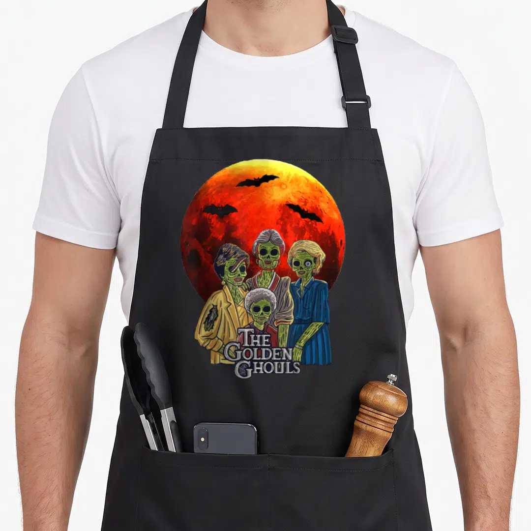 Ghoul golden apron - Four green zombie women under a red moon with three flying bats and stacked text — shown on a black cotton canvas apron against a white background