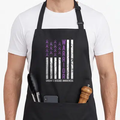 Funny warrior crohns awareness apron - Purple ribbons and white stripes with vertical WARRIOR text and distressed — shown on a black cotton canvas apron against a white background