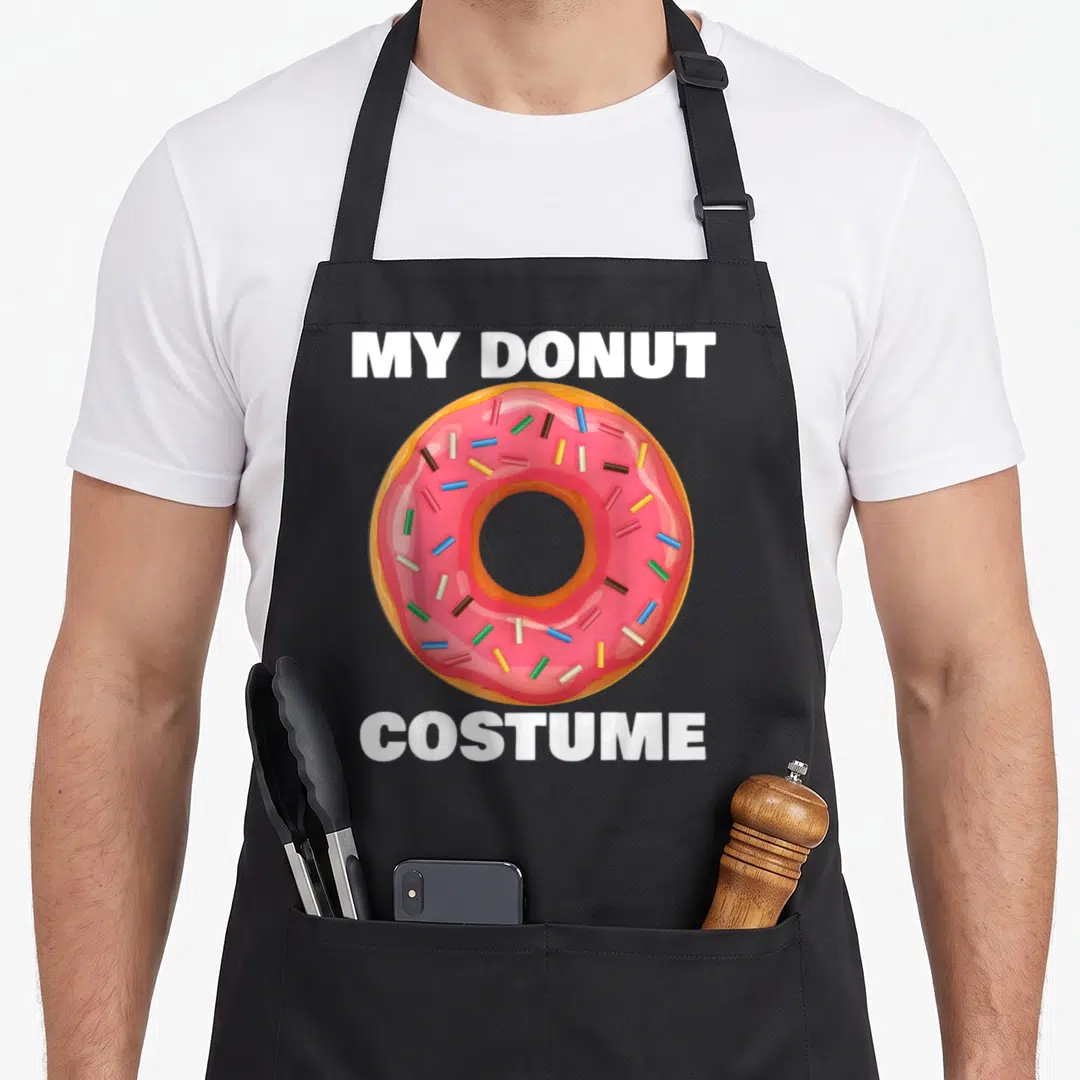 Halloween sprinkle apron - Cartoon pink donut with MY DONUT COSTUME text in white block letters — shown on a black cotton canvas apron against a white background