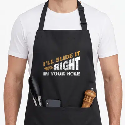 Slide hole apron - Orange and white distressed text I'll Slide It Right In Your Hole with small cornhole bag graphic — shown on a black cotton canvas apron against a white background