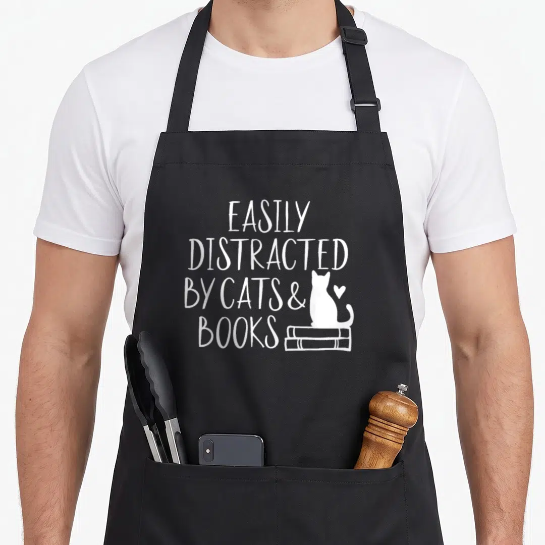 Distracted cat apron - White text EASILY DISTRACTED BY CATS & BOOKS next to cat on stacked books — shown on a black cotton canvas apron against a white background