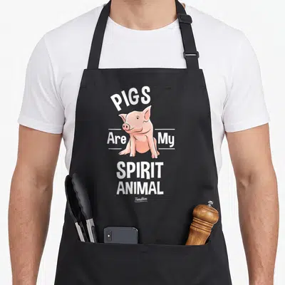 Cute pig animal apron - Cartoon pink pig sitting below arched PIGS text and stacked SPIRIT ANIMAL words — shown on a black cotton canvas apron against a white background