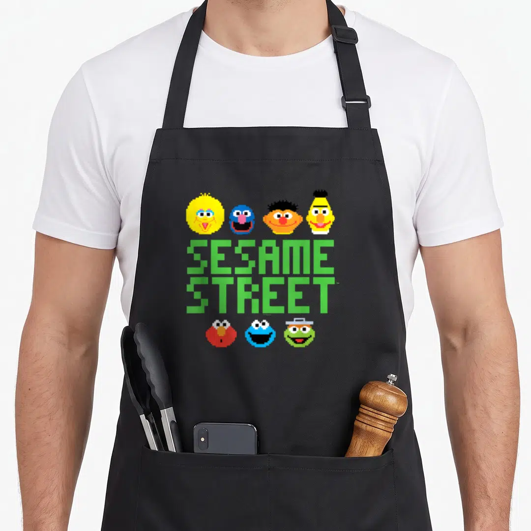 Cute pixel apron - Seven pixelated character heads surrounding stacked green SESAME STREET text — shown on a black cotton canvas apron against a white background