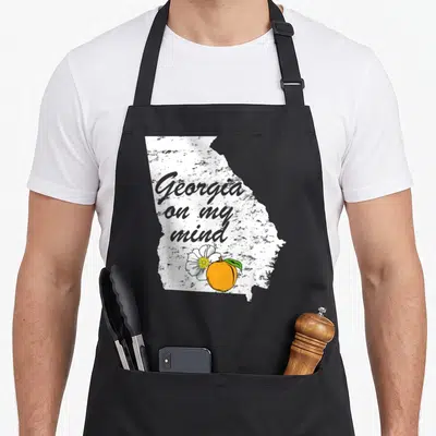 Mind southern aprons - Distressed white Georgia map with grey text, orange peach, and white flower — shown on a black cotton canvas apron against a white background
