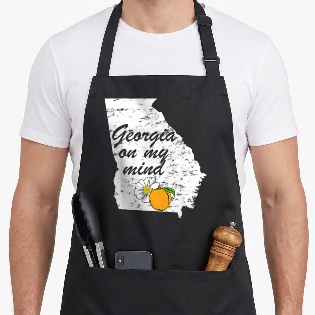 Mind southern aprons - Distressed white Georgia map with grey text, orange peach, and white flower — shown on a black cotton canvas apron against a white background