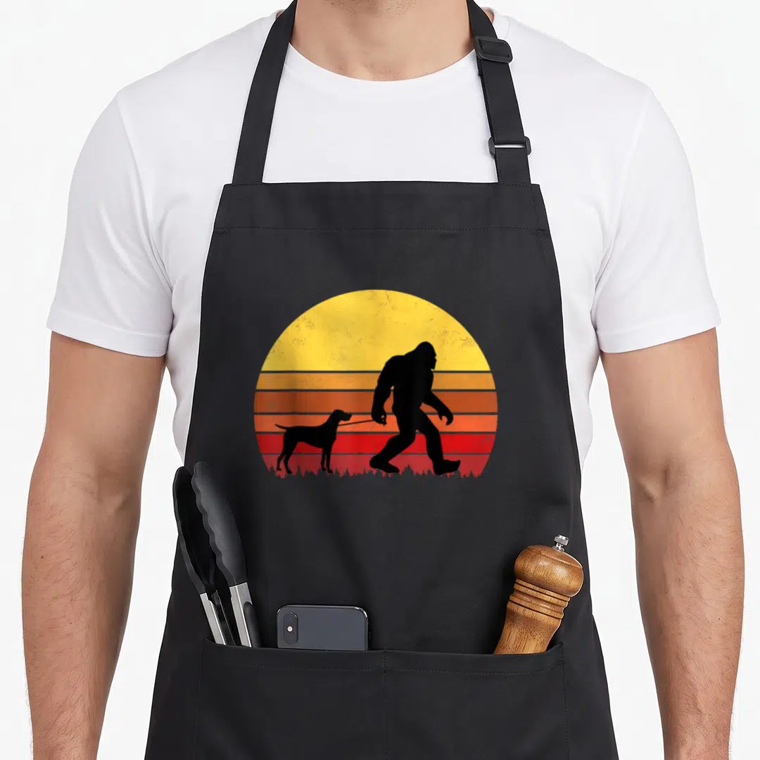 Shorthaired dog apron - Bigfoot silhouette walking a slender dog on a leash against a yellow orange and red striped sun — shown on a black cotton canvas apron against a white background